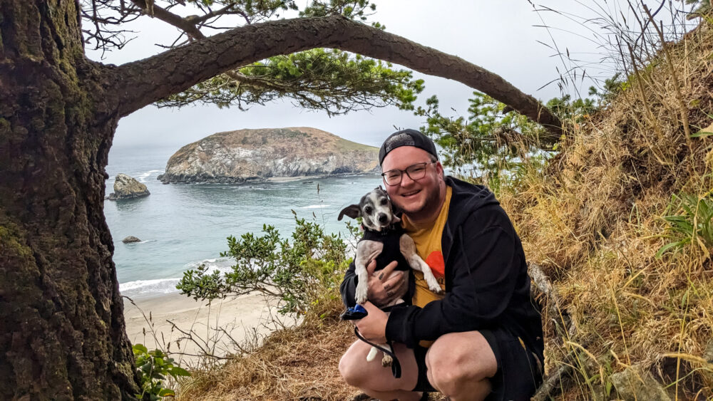 Miles crouched down on some rocks holding their dog, with a sweeping view of the ocean in the background, surrounded by some trees.