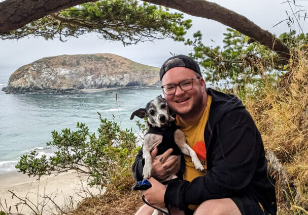 Miles crouched down on some rocks holding their dog, with a sweeping view of the ocean in the background, surrounded by some trees.